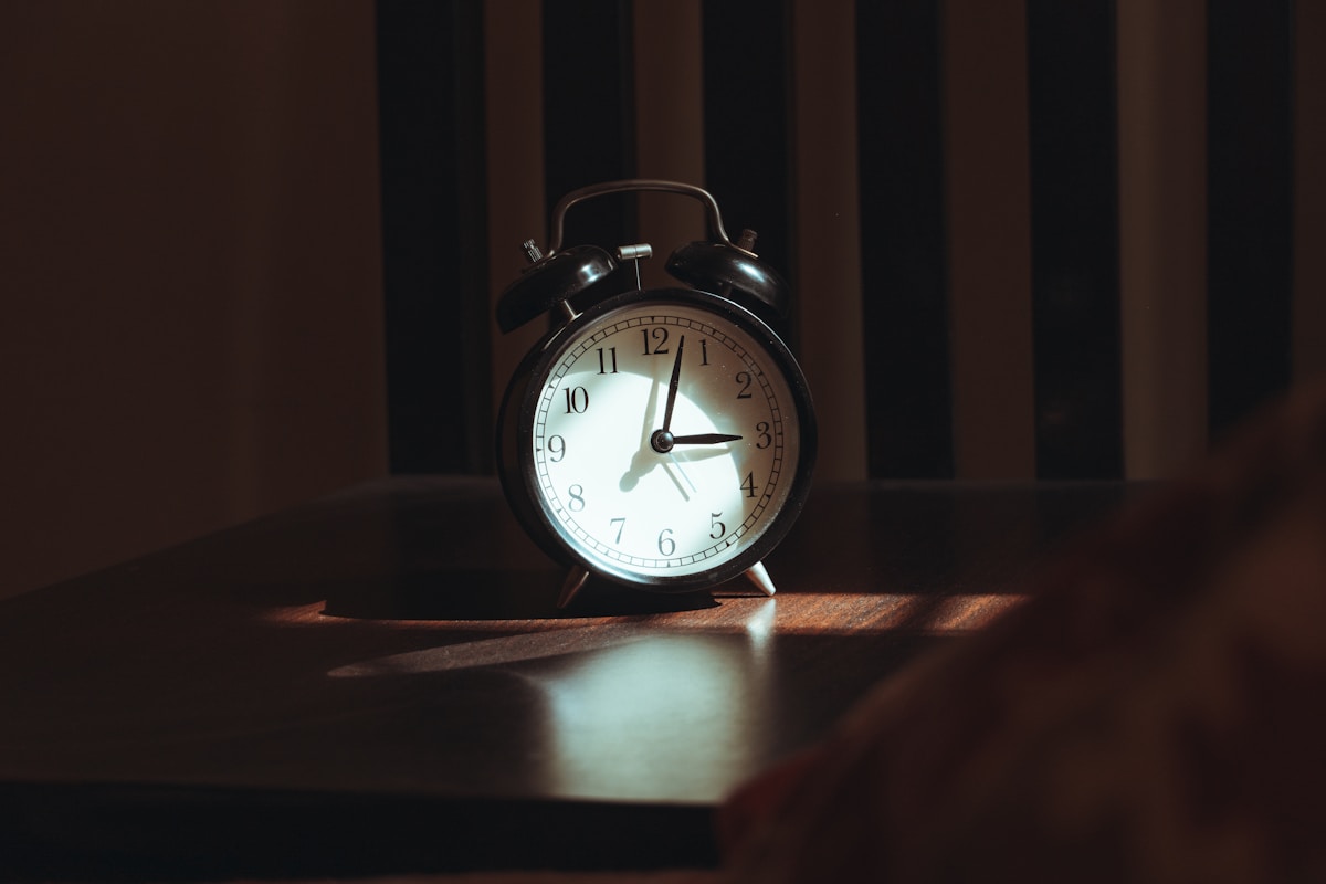 A black twin-bell analogue alarm clock on a dark wooden bedside table, lit by a narrow shaft of light, its hands reading a little after three
