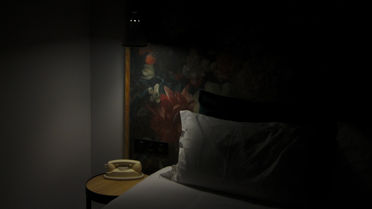 A dimly lit bedroom at night: a white pillow and bed in shadow, with a small round bedside table holding a cream rotary telephone under a wall lamp