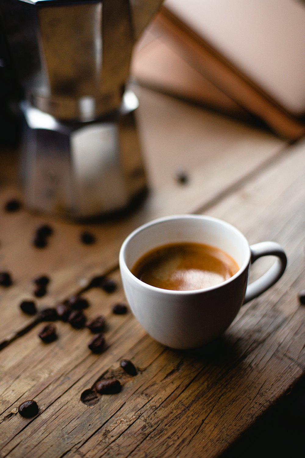 A white cup of espresso with a pale crema on a worn wooden table, roasted coffee beans scattered around it and a stovetop moka pot standing behind