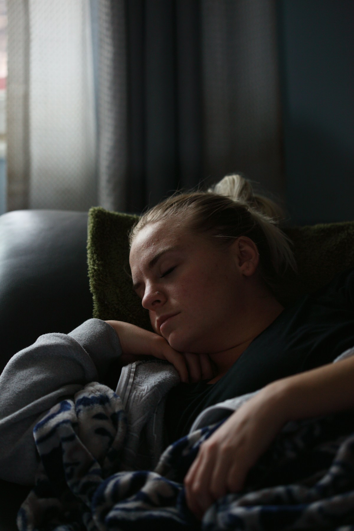 A woman asleep on a sofa in daylight, her head resting on her arm against a green cushion, wrapped in a patterned blanket